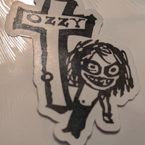 May include: A black and white sticker featuring a cartoon figure and a cross. The cross has the word "OZZY" printed on it. The cartoon figure has a wide grin and wild hair.