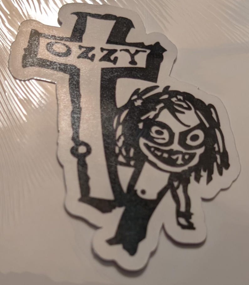 May include: A black and white sticker featuring a cartoon figure and a cross. The cross has the word "OZZY" printed on it. The cartoon figure has a wide grin and wild hair.