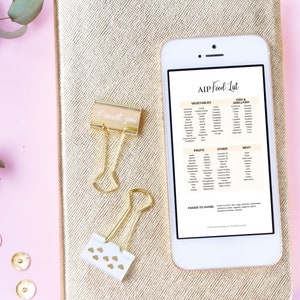 May include: A white smartphone displaying a list of foods allowed on the AIP diet. The list includes vegetables, fruits, meat, fish & shellfish, and other foods. The phone is on a gold notebook with a pink background.