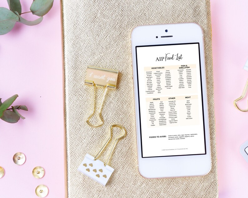 May include: A white smartphone displaying a list of foods allowed on the AIP diet. The list includes vegetables, fruits, meat, fish & shellfish, and other foods. The phone is on a gold notebook with a pink background.