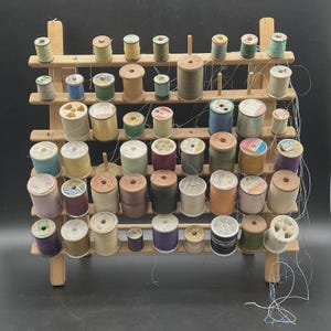 以下が含まれることがあります： さまざまな色の糸巻きが並んだ木製の糸巻きラック。糸巻きは複数の棚に配置され、一部の糸が垂れ下がっています。ラックは明るい色の木材で作られています。