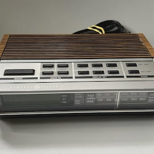 Puede incluir: Un radio reloj AM/FM General Electric con un panel superior de vetas de madera con listones horizontales, una interfaz de control plateada con botones negros y una ventana de visualización digital. Incluye un cable de alimentación negro con un lazo amarillo.