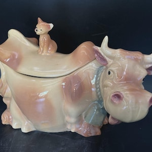 May include: A ceramic cow-shaped cookie jar with a glossy finish in shades of cream and light brown. The lid features a small cat figurine sitting on top. The cow has a blue collar with a bell around its neck and prominent horns on its head.