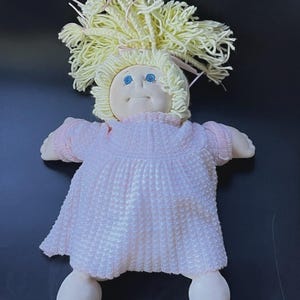 May include: A soft-sculpture doll with pale skin, blue eyes, and a small mouth. The doll has thick, light yellow yarn hair tied with small pink ribbons and wears a light pink, short-sleeved dress with a textured, ribbed knit pattern.