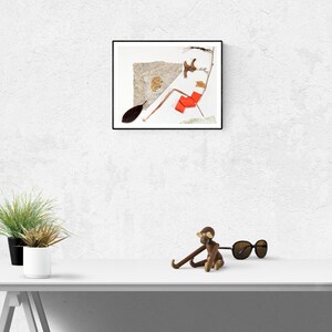 May include: A framed collage of dried leaves and twigs on a white background. A small wooden monkey figurine with sunglasses sits on a white table in front of the framed collage.