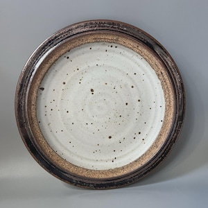 May include: A round ceramic plate featuring a speckled off-white center with concentric circular ridges. The plate has a wide, textured rim finished in a dark brown and bronze metallic glaze that transitions into a deep black edge.