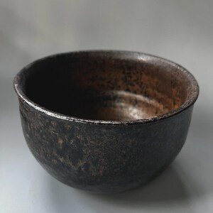 May include: A round ceramic bowl with a dark, textured exterior featuring mottled black and deep brown tones. The interior of the bowl displays a similar dark, weathered finish with a smooth, slightly reflective surface along the rim.