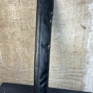 May include: A black leather sheath, likely for a long knife or sword, is shown against a neutral background. The sheath is long and narrow, with a curved shape and several rivets along the side. The leather appears to be slightly textured.