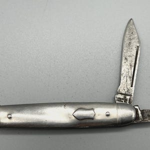 May include: A vintage pocket knife with a silver-colored handle and two blades. One blade is fully extended, and the other is partially open. The knife shows signs of wear, with a tarnished appearance.