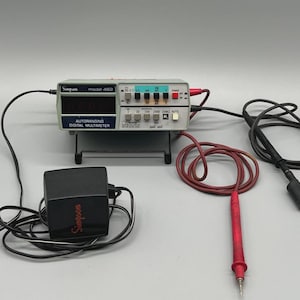 May include: A Simpson Model 462 autoranging digital multimeter with a black power adapter and red and black test leads. The multimeter has a digital display and various buttons and switches. The unit is on a small stand.