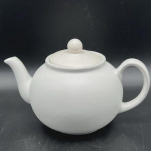 Puede incluir: Una tetera de cerámica blanca con cuerpo redondeado, boquilla curva y asa de bucle. La tapa tiene un pequeño pomo en la parte superior. La tetera está sobre un fondo oscuro.