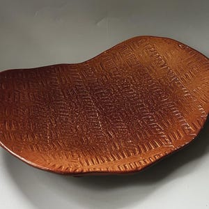 Könnte beinhalten: Ein oval geformtes Keramiktablett mit einer satten, rotbraunen Glasur. Die Oberfläche weist eine tief eingeprägte, sich wiederholende gitterartige Textur auf, die das gesamte Innere bedeckt und ein strukturiertes Aussehen auf der flachen Schale erzeugt.