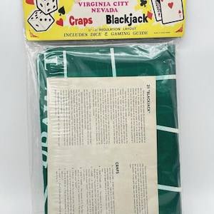 May include: A packaged gaming layout for Craps and Blackjack, featuring a green felt surface with white lines. The packaging includes dice, playing cards, and a gaming guide. The text on the packaging reads "Reno Tahoe Virginia City Nevada".
