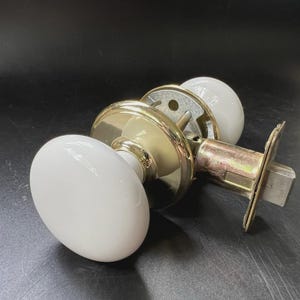 Puede incluir: Un juego de pomos para puerta con dos tiradores redondos y lisos de porcelana blanca unidos a rosetas circulares de latón pulido. El mecanismo incluye un pestillo metálico con una placa frontal rectangular de diseño clásico.