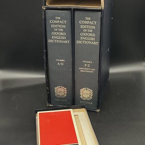 Könnte beinhalten: Ein Satz von The Compact Edition of the Oxford English Dictionary, Bände I und II, in einem dunkelblauen Etui. Eine Leseglas mit rotem Deckel und dem Text "Reading Glass" befindet sich in einer separaten Box.