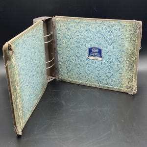 May include: An open vintage binder with a metal spine and a blue and white patterned interior. The cover is a faded blue-green with a distressed look. A label with text is visible on the inside cover.