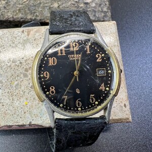 Puede incluir: Un reloj Citizen Quartz con esfera negra, detalles dorados y visualización de la fecha a las 3. El reloj tiene una caja plateada y una correa de cuero negro. La esfera muestra números y agujas doradas.