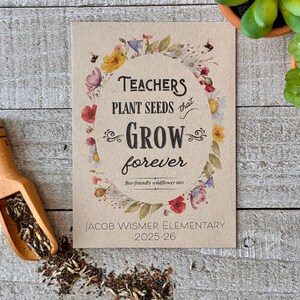 May include: A seed packet with the text "TEACHERS PLANT SEEDS that GROW forever" surrounded by a floral wreath. The packet also includes "Bee-friendly wildflower mix" and "JACOB WISMER ELEMENTARY 2025-26". A wooden scoop and seeds are next to the packet.