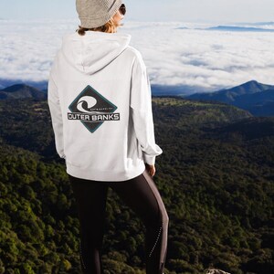 Peut inclure: Un sweat-shirt à capuche blanc avec un motif en forme de losange dans le dos. Le graphisme présente une icône de vague stylisée, le texte "NORTH CAROLINA" en petites lettres et les mots "OUTER BANKS" dans un cadre rectangulaire noir.
