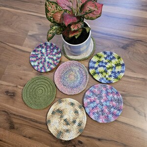 May include: A collection of six circular crochet coasters in various colors including solid olive green, variegated pink and blue, blue and green, and neutral earth tones, arranged around a potted plant with pink and green variegated leaves.