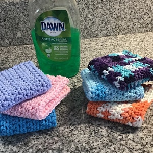 May include: A stack of handmade crochet washcloths in various colors, including blue, pink, purple, and orange. A bottle of Dawn Ultra Antibacterial dish soap is in the background. The washcloths are square and have a textured appearance.