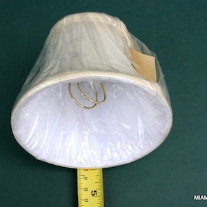 May include: A white lampshade with a gold metal harp, measuring 5 inches in diameter.