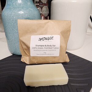 以下が含まれることがあります： 白いラベルに「INDULGE BATH AND BODY」と「Shampoo & Body Bar 100% Grass-Fed Beef Tallow Rosemary & Lavender Essential Oil 3 oz www.indulgebathandbody.com」と書かれた茶色の紙袋。白い石鹸が黒い表面の上に置かれている。