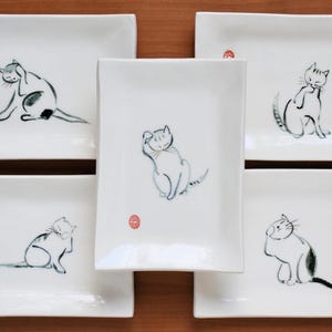 May include: A set of five rectangular white ceramic plates, each featuring a unique black ink-style illustration of a cat in a different pose. Each plate includes a small, circular red stamp mark near the cat illustration on the white surface.