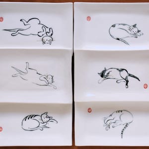 May include: A set of six rectangular white ceramic plates, each featuring a unique black ink-style illustration of a cat in a different resting pose. Each plate includes a small, circular red stamp mark near the edge of the design.