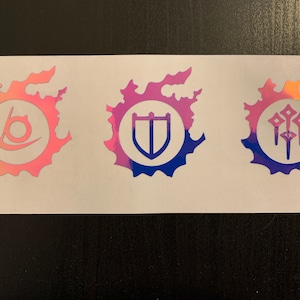 Peut inclure: Trois autocollants irisés sur fond blanc. Chaque autocollant présente un symbole différent à l'intérieur d'un motif circulaire à bords dentelés. Le premier autocollant est rose et présente une ligne courbe avec un cercle à l'intérieur. Le deuxième autocollant est violet et bleu et présente un bouclier. Le troisième autocollant est violet et orange et présente trois lances.