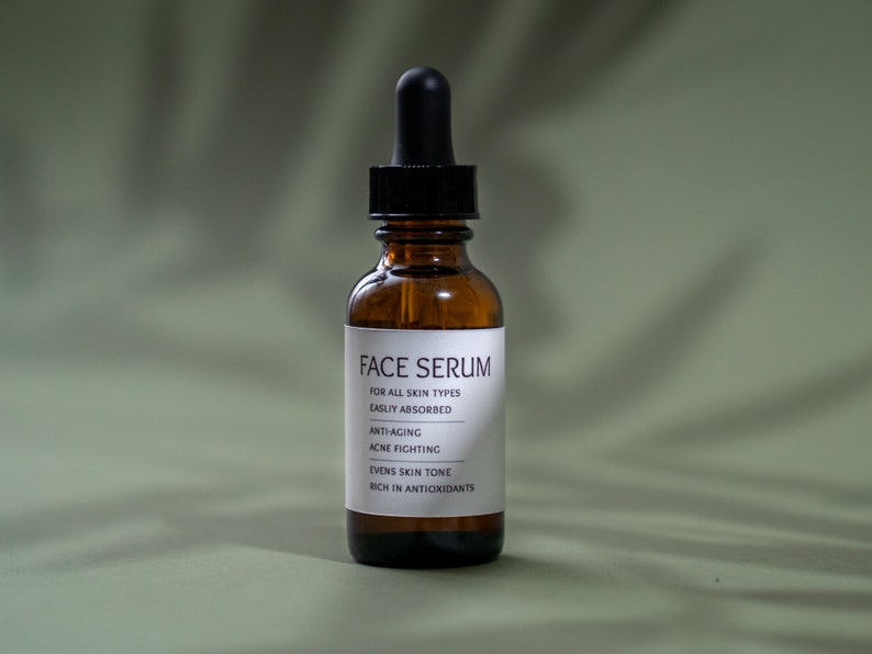 Op de afbeelding: Een bruine glazen fles met een druppelaar, met het label "FACE SERUM" en een opsomming van de voordelen: voor alle huidtypen, gemakkelijk te absorberen, anti-aging, acne-bestrijdend, egaliseert de teint, rijk aan antioxidanten.