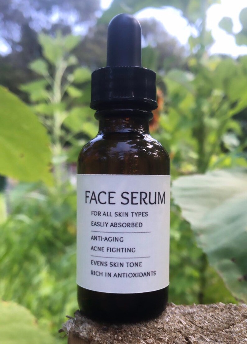 Op de afbeelding: Een bruine glazen fles met een zwarte druppelaar en een wit etiket met de tekst "FACE SERUM" en de voordelen: "VOOR ALLE HUIDTYPES GEMAKKELIJK OPGENOMEN ANTI-VEROUDERING ACNE BESTRIJDING VERZACHT DE HUIDTOON RIJKE AANTAL ANTIOXIDANTEN".