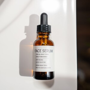 Op de afbeelding: Een bruine glazen fles met een druppelaar en een wit etiket waarop "FACE SERUM" staat en de voordelen van het serum worden vermeld: "VOOR ALLE HUIDTYPES", "GEMAKKElijk OPGENOMEN", "ANTI-AGING", "ACN&Eacute; BESTRIJDING", "VEREVEELT DE HUIDTOON", "RIJK AAN ANTIOXIDANTEN".