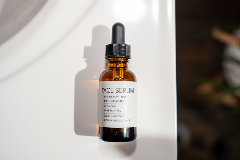 Op de afbeelding: Een bruine glazen fles met een druppelaar en een wit etiket waarop "FACE SERUM" staat en de voordelen van het serum worden vermeld: "VOOR ALLE HUIDTYPES", "GEMAKKElijk OPGENOMEN", "ANTI-AGING", "ACN&Eacute; BESTRIJDING", "VEREVEELT DE HUIDTOON", "RIJK AAN ANTIOXIDANTEN".