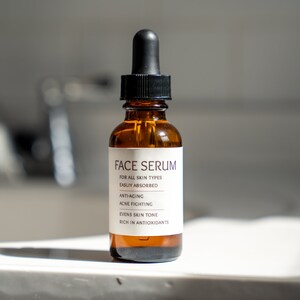 Op de afbeelding: Een bruine glazen fles met een zwarte druppelaar en een wit etiket waarop "FACE SERUM" staat en de voordelen van het serum worden vermeld: "VOOR ALLE HUIDTYPES", "GEMAKKElijk OPGENOMEN", "ANTI-AGING", "ACNE BESTRIJDEND", "VEREVEELT DE HUIDTOON" en "RIJK AAN ANTIOXIDANTEN".