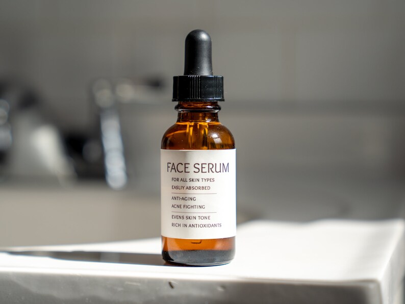 Op de afbeelding: Een bruine glazen fles met een zwarte druppelaar en een wit etiket waarop "FACE SERUM" staat en de voordelen van het serum worden vermeld: "VOOR ALLE HUIDTYPES", "GEMAKKElijk OPGENOMEN", "ANTI-AGING", "ACNE BESTRIJDEND", "VEREVEELT DE HUIDTOON" en "RIJK AAN ANTIOXIDANTEN".