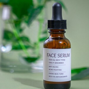 Op de afbeelding: Een bruine glazen fles met een zwarte druppelaar, met het label "FACE SERUM". Het label vermeldt ook de voordelen van het product: "FOR ALL SKIN TYPES", "EASILY ABSORBED", "ANTI-AGING", "ACNE FIGHTING", "EVENS SKIN TONE" en "RICH IN ANTIOXIDANTS".