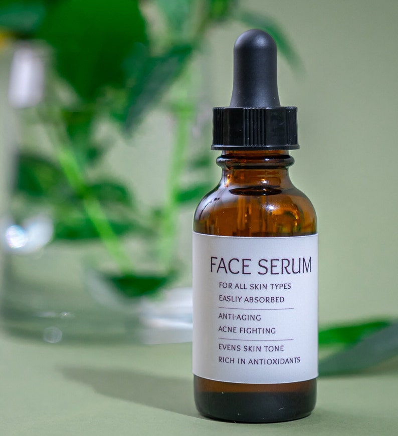 Op de afbeelding: Een bruine glazen fles met een zwarte druppelaar, met het label "FACE SERUM". Het label vermeldt ook de voordelen van het product: "FOR ALL SKIN TYPES", "EASILY ABSORBED", "ANTI-AGING", "ACNE FIGHTING", "EVENS SKIN TONE" en "RICH IN ANTIOXIDANTS".