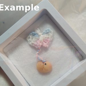 May include: A small, crocheted butterfly with pink, blue, and white yarn, hanging from a white string. The butterfly is attached to a small, peach-colored, clay figurine.
