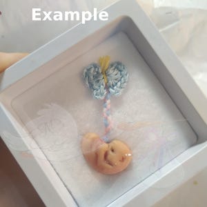 以下が含まれることがあります： ガラスの前面を持つ小さな白い箱に、手作りの桃色の豆が、青と黄色の蝶が上に置かれた状態で展示されています。蝶は糸でできており、豆にピンクと青の紐が付けられています。
