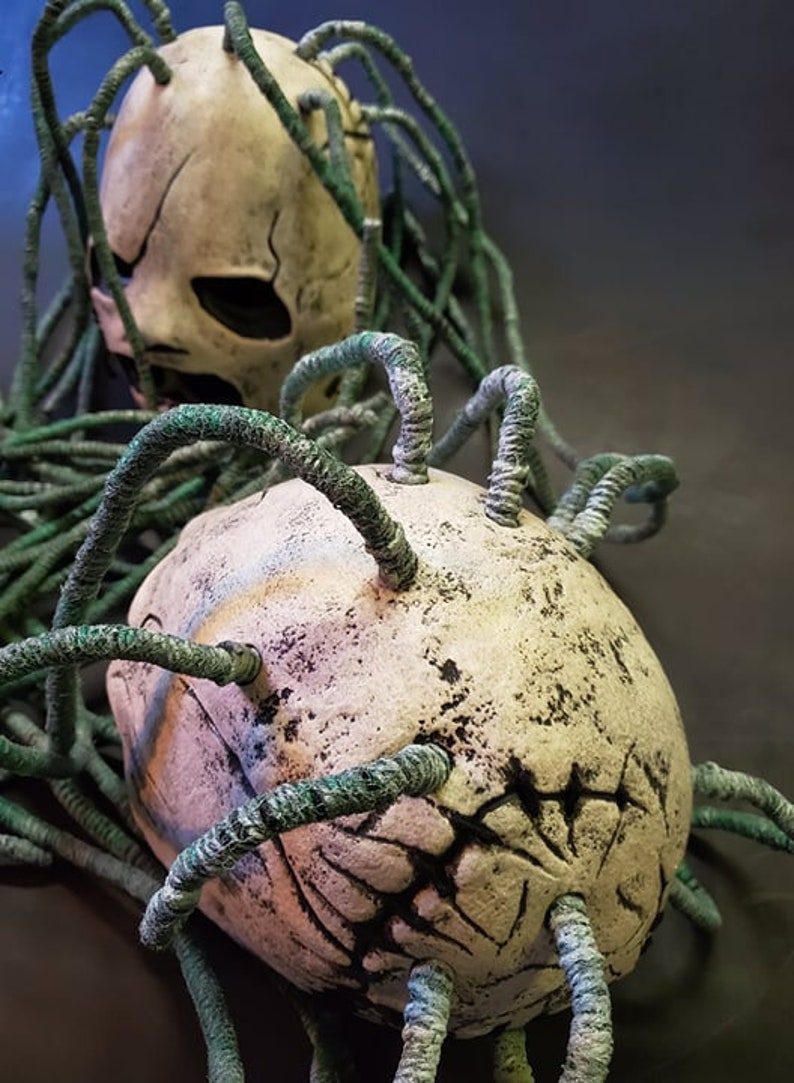 Peut inclure: Deux cr&acirc;nes en c&eacute;ramique blanche avec des vrilles vertes, ressemblant &agrave; une plante parasite, qui poussent de leur t&ecirc;te. Les cr&acirc;nes ont un aspect vieilli et d&eacute;labr&eacute; avec des marques noires.