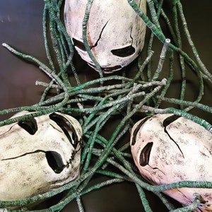 Peut inclure: Trois masques de cr&acirc;ne blancs avec des yeux et des bouches noirs, recouverts de ficelle verte. Les masques ont un aspect vieilli et d&eacute;labr&eacute;.