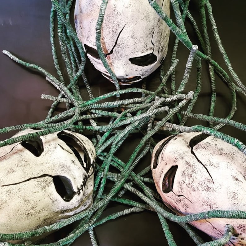 Peut inclure: Trois masques de cr&acirc;ne blancs avec des yeux et des bouches noirs, recouverts de ficelle verte. Les masques ont un aspect vieilli et d&eacute;labr&eacute;.