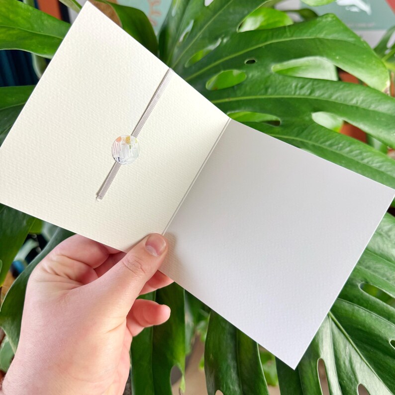 Pode incluir: Um cart&atilde;o branco em branco com um pequeno adesivo redondo e colorido na frente. O cart&atilde;o est&aacute; dobrado ao meio e segurado por uma m&atilde;o.