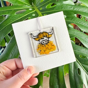 Pode incluir: Um ornamento de vidro com uma ilustra&ccedil;&atilde;o em preto e branco de uma vaca das terras altas com uma juba texturizada amarela e laranja. O ornamento est&aacute; pendurado em um cord&atilde;o branco e &eacute; exibido em um cart&atilde;o branco.