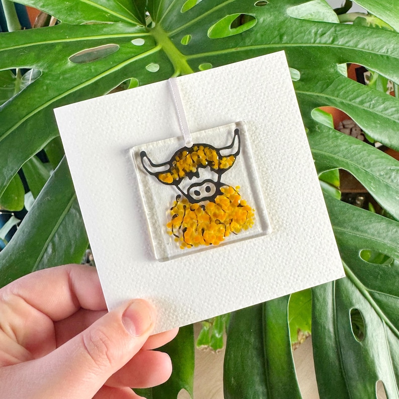 Pode incluir: Um ornamento de vidro com uma ilustra&ccedil;&atilde;o em preto e branco de uma vaca das terras altas com uma juba texturizada amarela e laranja. O ornamento est&aacute; pendurado em um cord&atilde;o branco e &eacute; exibido em um cart&atilde;o branco.