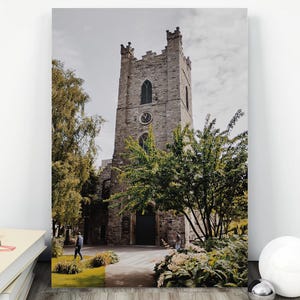 May include: A vertical rectangular wall art print featuring a stone church tower with a clock face, arched windows, and crenelated top. The scene includes green trees, a paved path, and a person walking near the entrance under a cloudy sky.