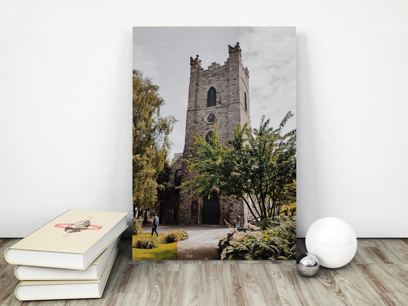 May include: A vertical rectangular wall art print featuring a stone church tower with a clock face, arched windows, and crenelated top. The scene includes green trees, a paved path, and a person walking near the entrance under a cloudy sky.
