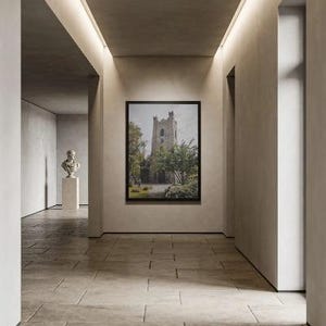 May include: A framed wall art print depicting a stone church tower surrounded by green trees and foliage, displayed on a neutral-toned wall within a hallway. A white bust sculpture on a pedestal is visible in the background of the corridor.