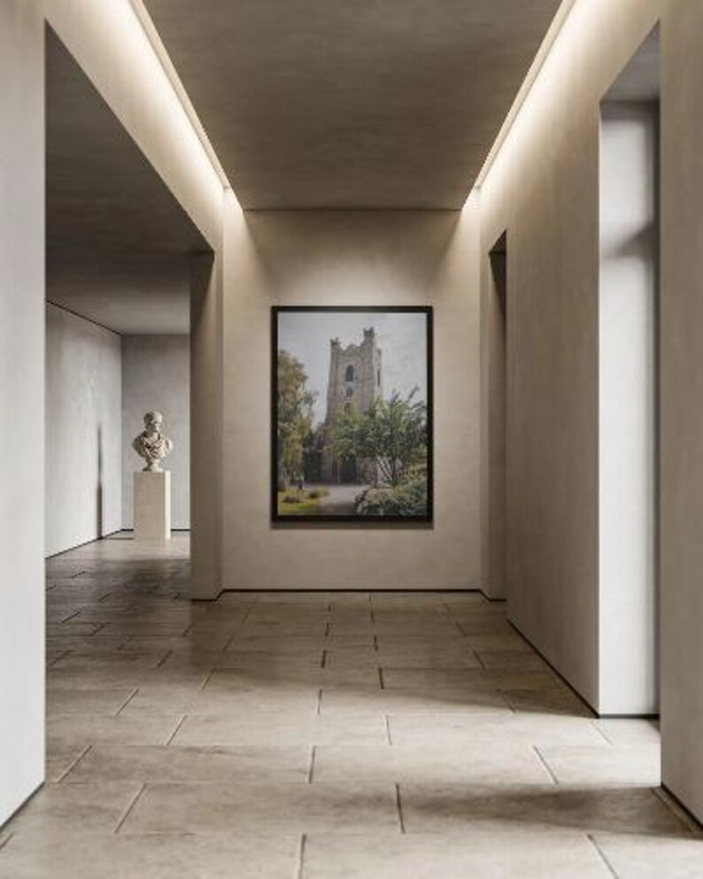 May include: A framed wall art print depicting a stone church tower surrounded by green trees and foliage, displayed on a neutral-toned wall within a hallway. A white bust sculpture on a pedestal is visible in the background of the corridor.